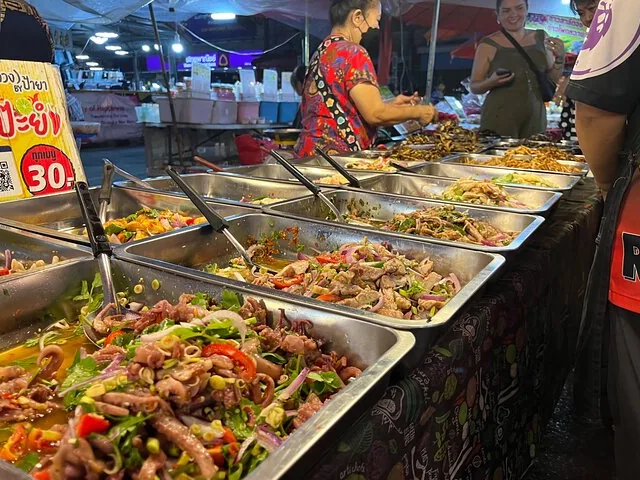Street food stall with metal trays full of Thai salads and vendors.
